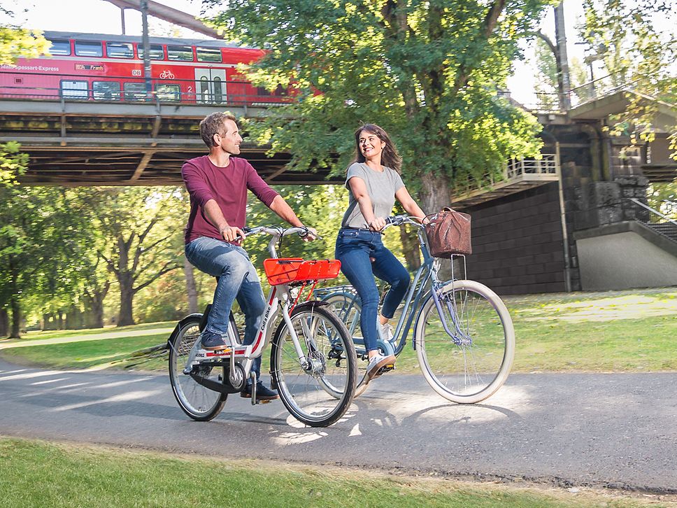 Pärchen fährt auf privatem Rad und Call a Bike durch einen Park. Im Hintergrund eine Regionalbahn von DB Regio.