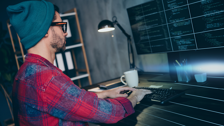 Mann mit Mütze und Brille an Schreibtisch sitzend vor Bildschirmen mit Code