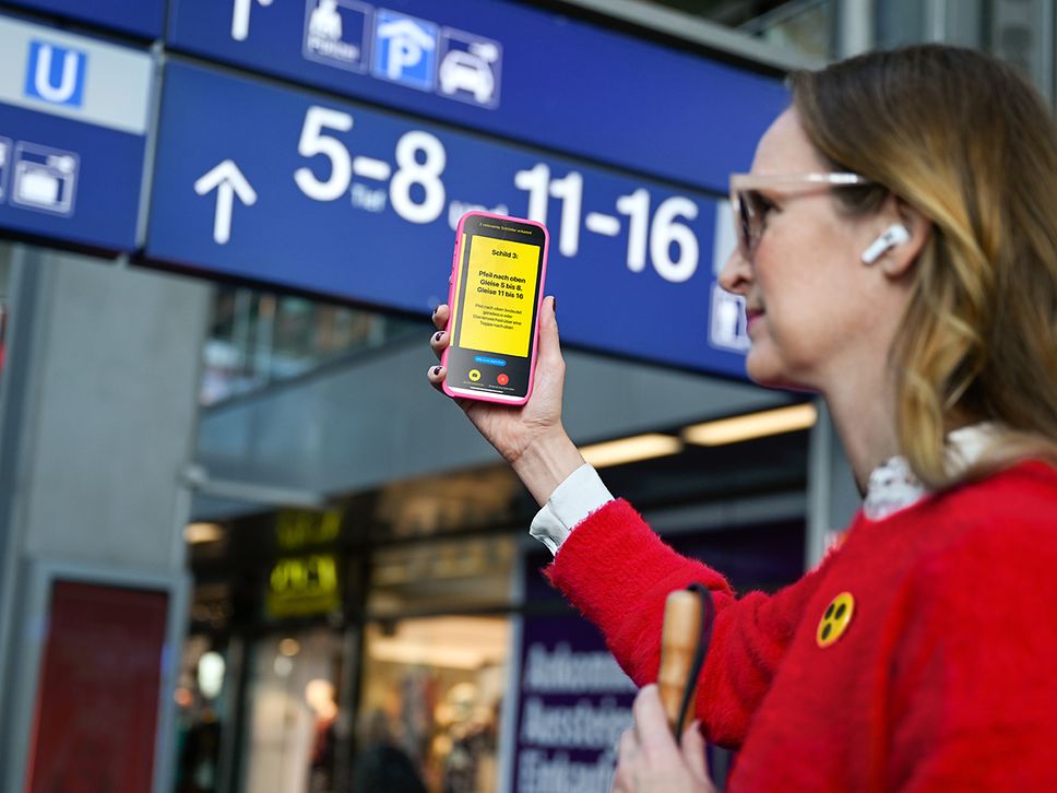 Eine Frau steht am Bahnhof mit einem Handy in der Hand. Die App zum Barrierefreien Reisen ist geöffnet und liest ihr Infos vor.