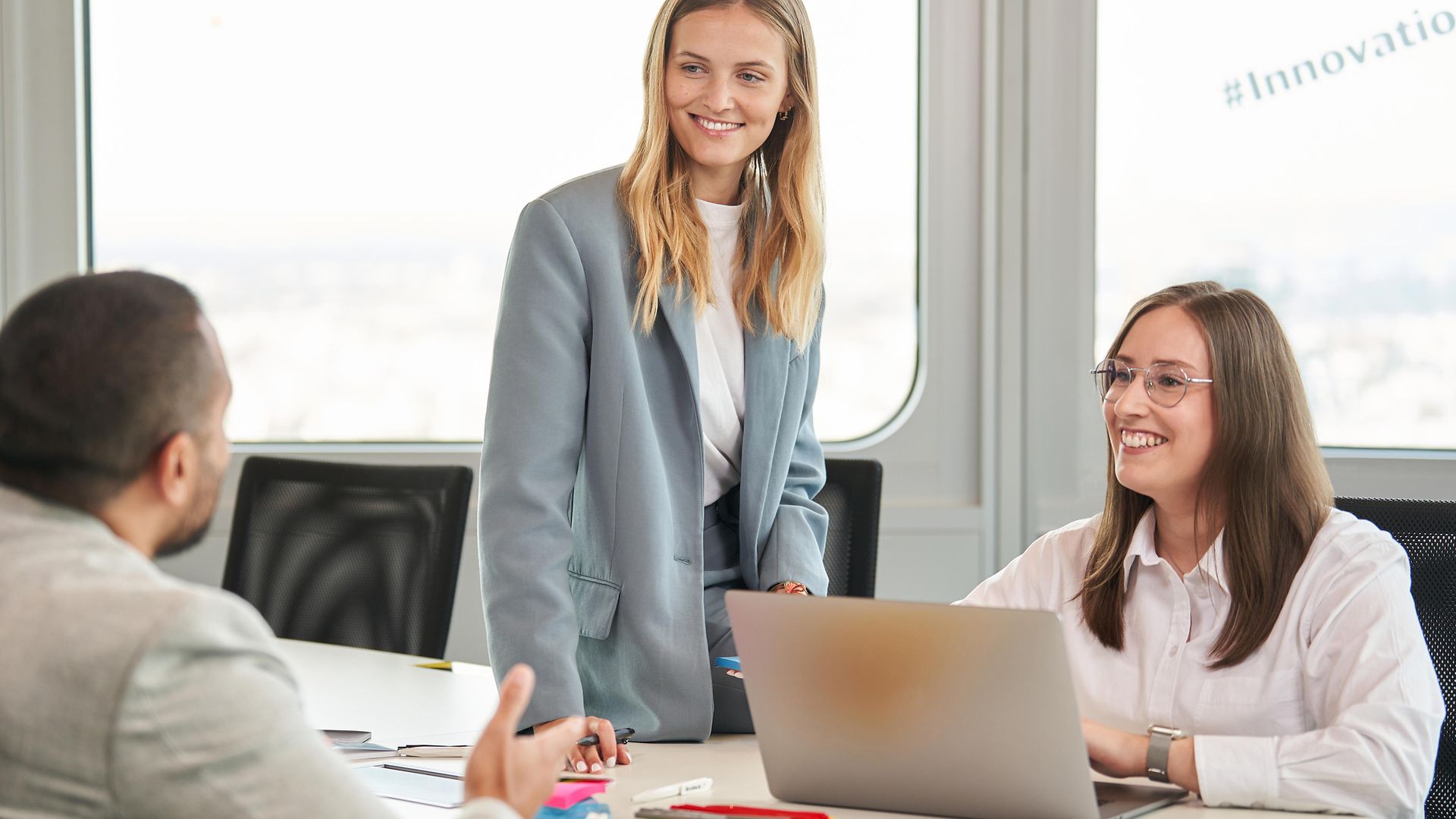 Zwei Frauen, freudig, im Gespräch mit einem Mann in einem Büro vor einem Laptop.