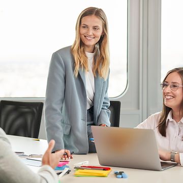 Zwei Frauen, freudig, im Gespräch mit einem Mann in einem Büro vor einem Laptop.