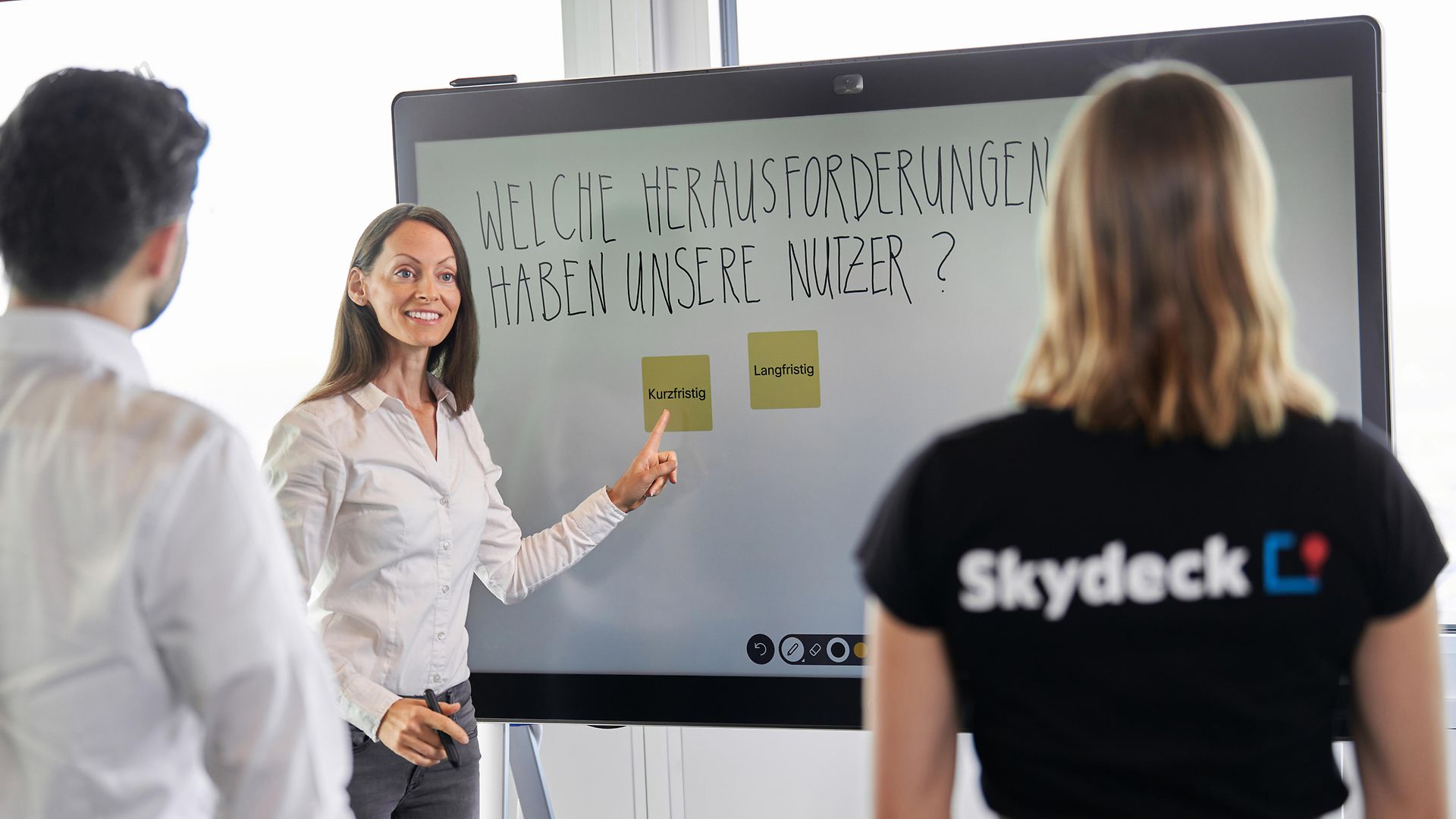 Frau stehend vor großem Screen, Mann und Frau davor hören zu