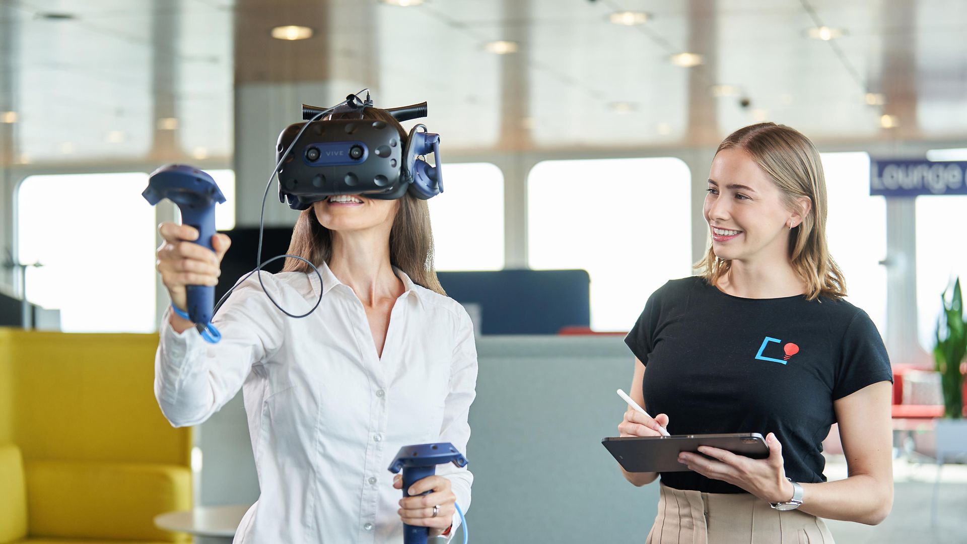 Frau mit VR-Brille, daneben zweite Frau freundlich grinsend mit Tablet