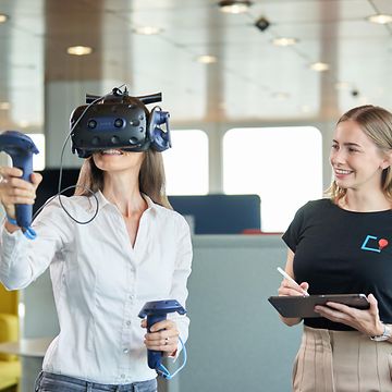 Woman with VR glasses, next to her a second woman with a tablet and a friendly grin.